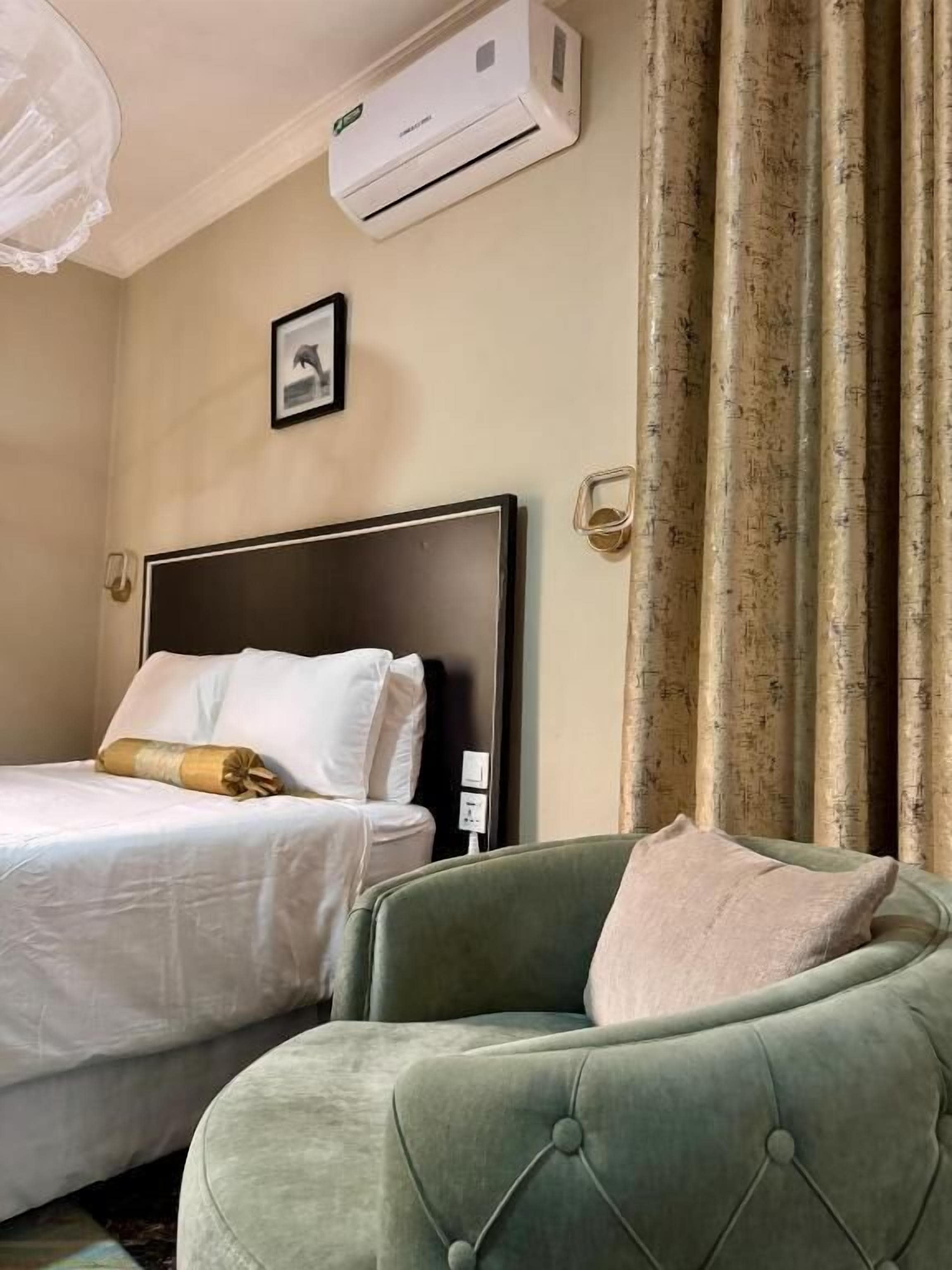 Una habitación de hotel con una cama, un sillón verde, un equipo de aire acondicionado, un cuadro en la pared y una cortina.