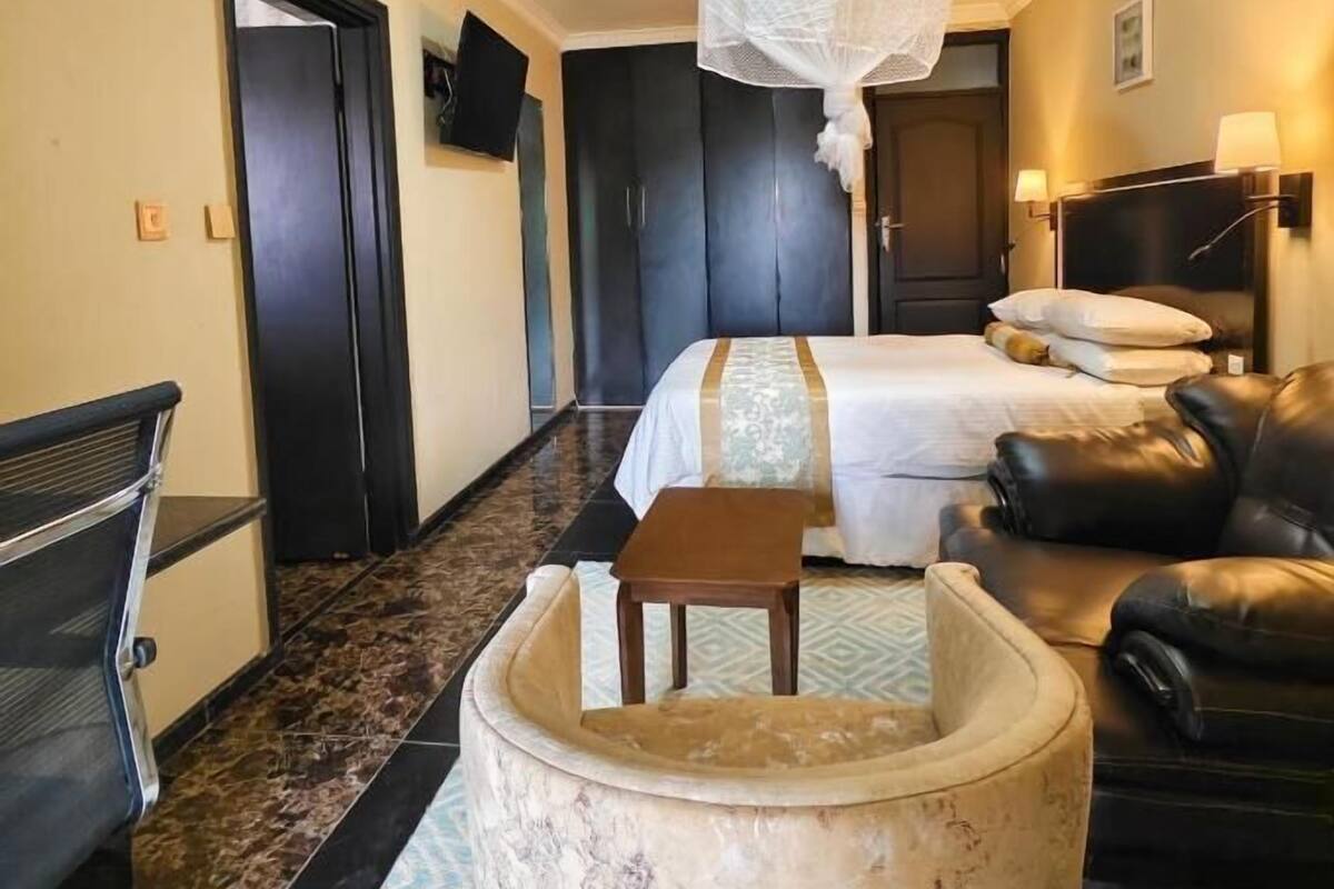 Una habitación de hotel con una cama, un sofá de cuero, una mesita redonda pequeña, un televisor y una mosquitera.