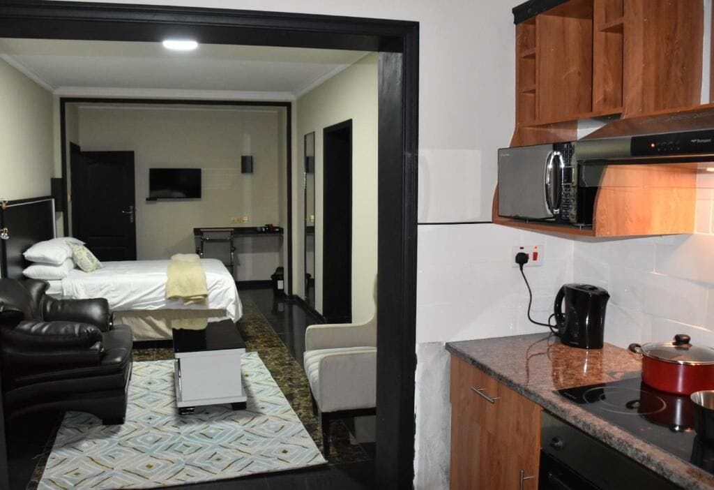 Una habitación de hotel compacta con una cama, zona de estar, kitchenette con microondas, cafetera y una olla roja.