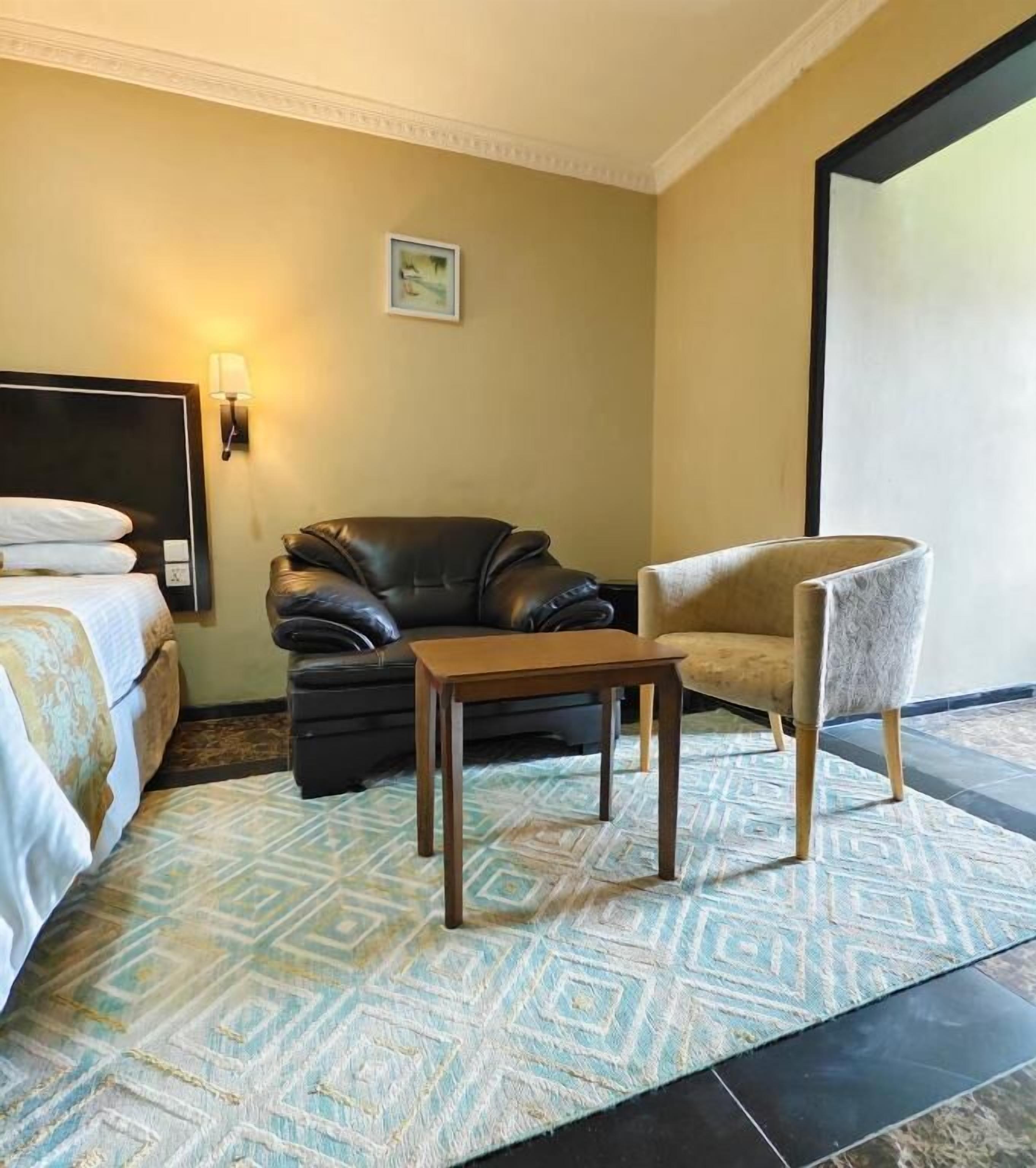 Ett hotellrum med en säng, en soffa i läder, ett träbord, en ljusfärgad fåtölj och en mönstrad matta.