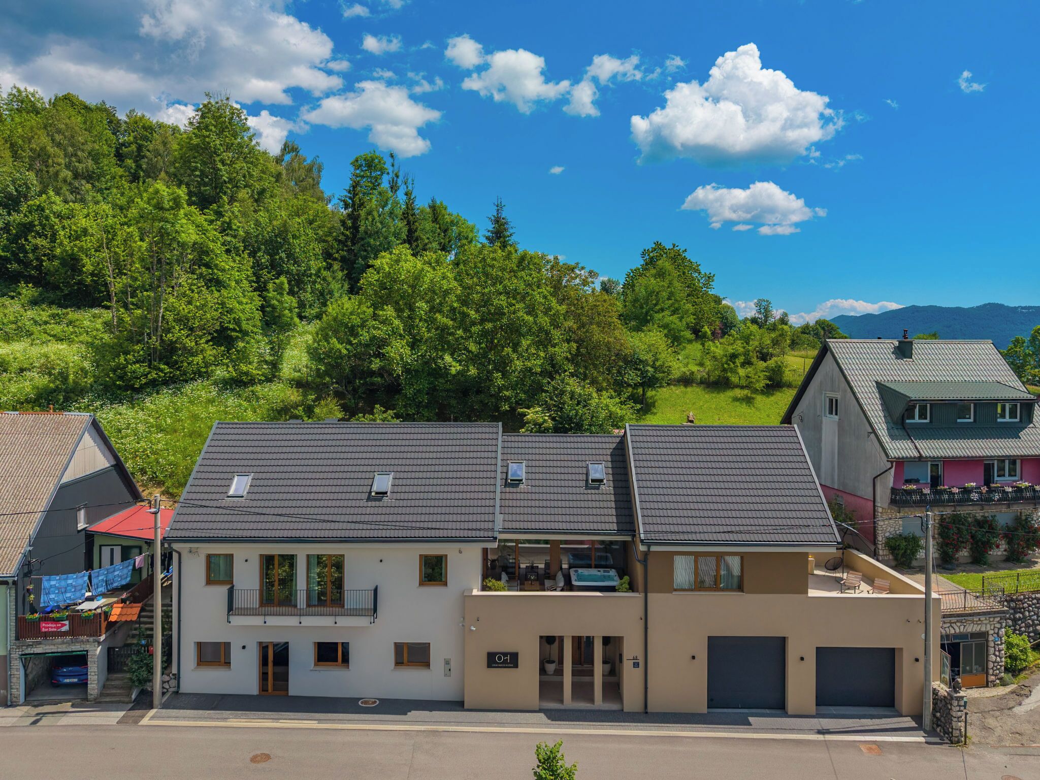 Ett bostadsområde med hus som har sluttande tak, en central byggnad med en balkong och en bakgrund av frodiga, gröna träd och kullar.