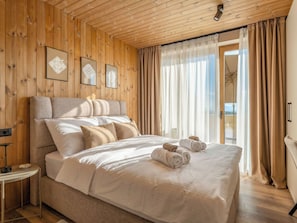 Ein Schlafzimmer mit Holzbalkendecke, einem Bett mit weißer Bettwäsche und Blick ins Freie durch große Fenster.
