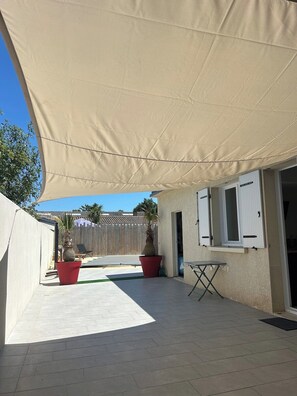 A covered patio area with a beige shade sail, two potted plants, a folding table, and a door with shutters.