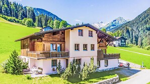 A two-story chalet with a balcony, surrounded by a stone wall, set in a mountainous area with green fields.
