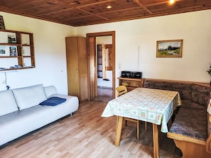 Een kamer met een houten plafond, een witte bank, een eettafel met tafelkleed, een houten bankje en een schilderij aan de muur.