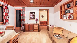 A cozy living room with a wooden ceiling, a dining area with a table and chairs, a sofa with cushions, and a bookshelf.
