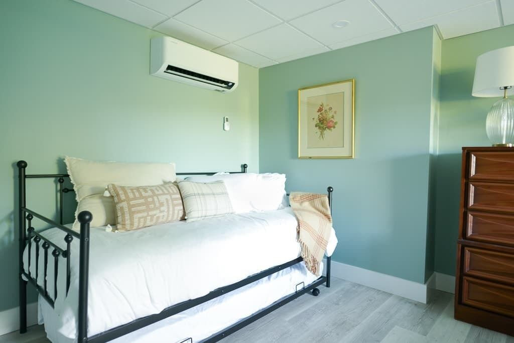 Una cama con colchón blanco y varias almohadas, un aparador de madera, un cuadro enmarcado en la pared y un equipo de aire acondicionado.