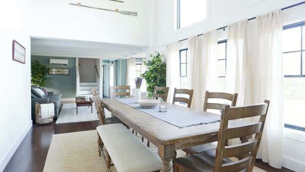 Een eethoek met een lange houten tafel, zes stoelen en een bank. Er zijn grote ramen met gordijnen, een plant en uitzicht op een andere kamer.