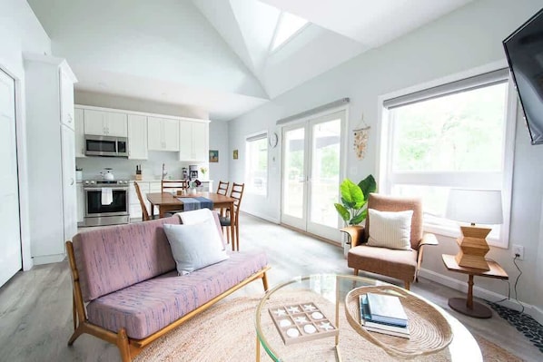 A modern kitchen with white cabinets, a dining area with a table and chairs, a sofa, and a coffee table with books.