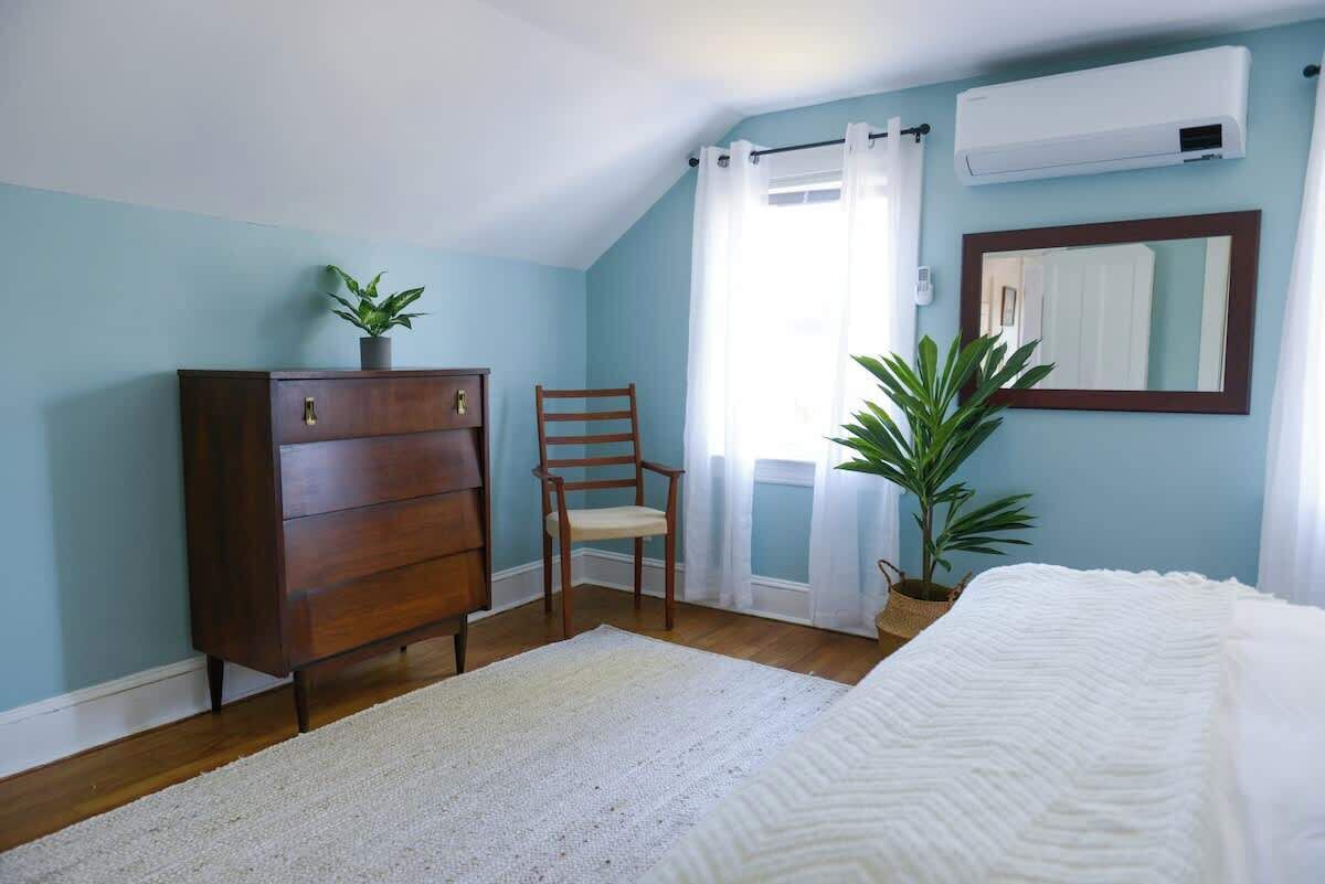 Ein Schlafzimmer mit einem Holzkapstativ, einem Sessel, einem Bett, einer Topfpflanze, einem Spiegel und einer Klimaanlage.