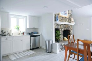 A modern kitchen with white cabinets, a dishwasher, a stone fireplace, and a dining area with a wooden table and chairs. - Fully Renovated "Saltbox" Home in Boothbay (Boothbay)