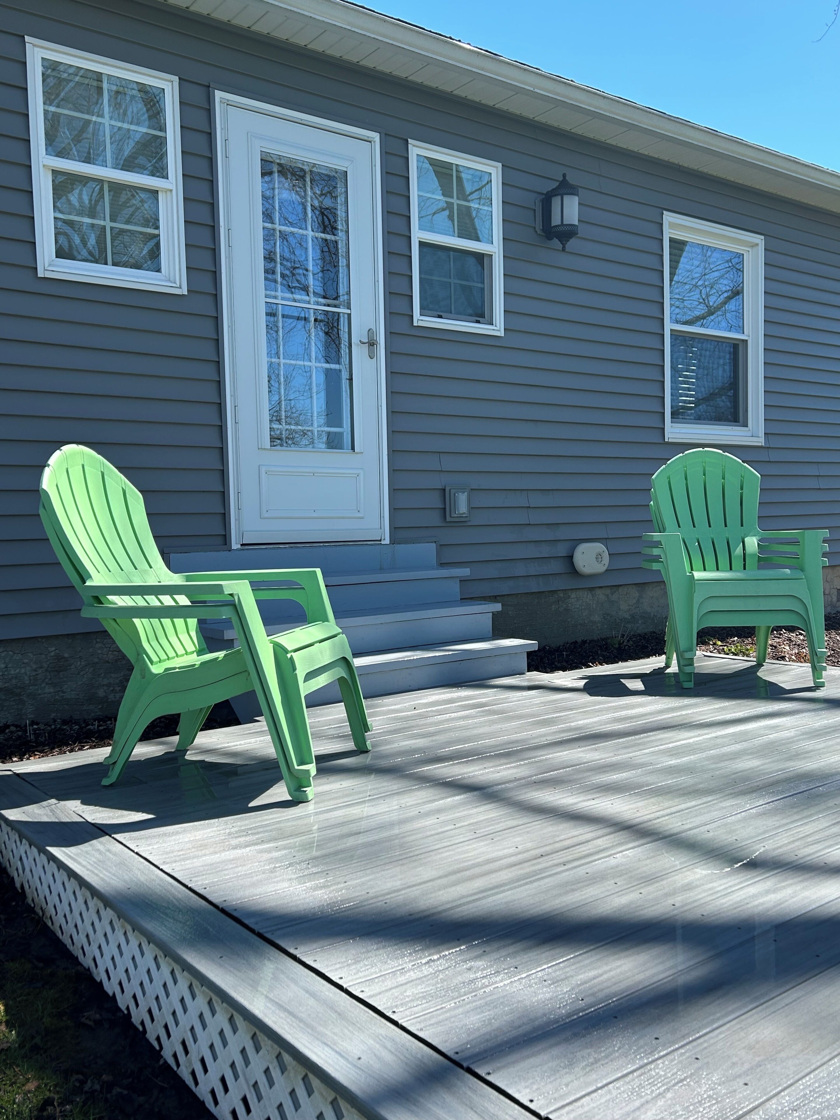 A house with two green Adirondack chairs on a wooden deck, a white front door, and a small window above the door.