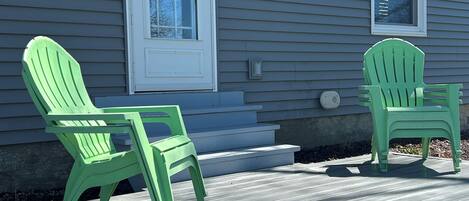 A house with two green Adirondack chairs on a wooden deck, a white front door, and a small window above the door.