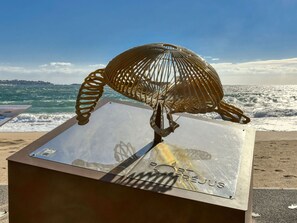Eine Metallskulptur eines Vogels auf einer spiegelnden Fläche mit Küstenlandschaft im Hintergrund.