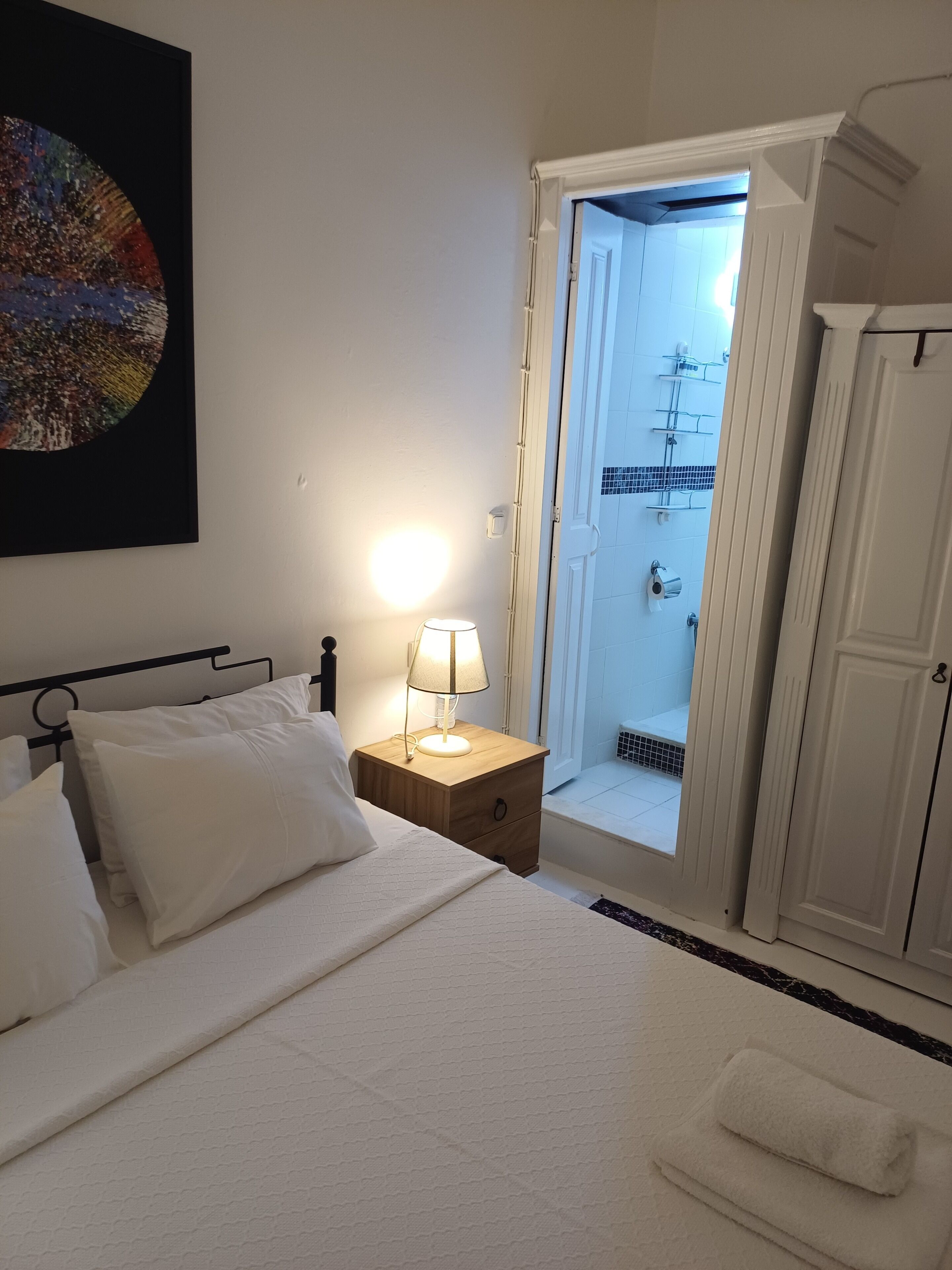 Habitación de hotel con cama, lámpara de noche, mesita de noche, baño con ducha y puerta que da a otra habitación.