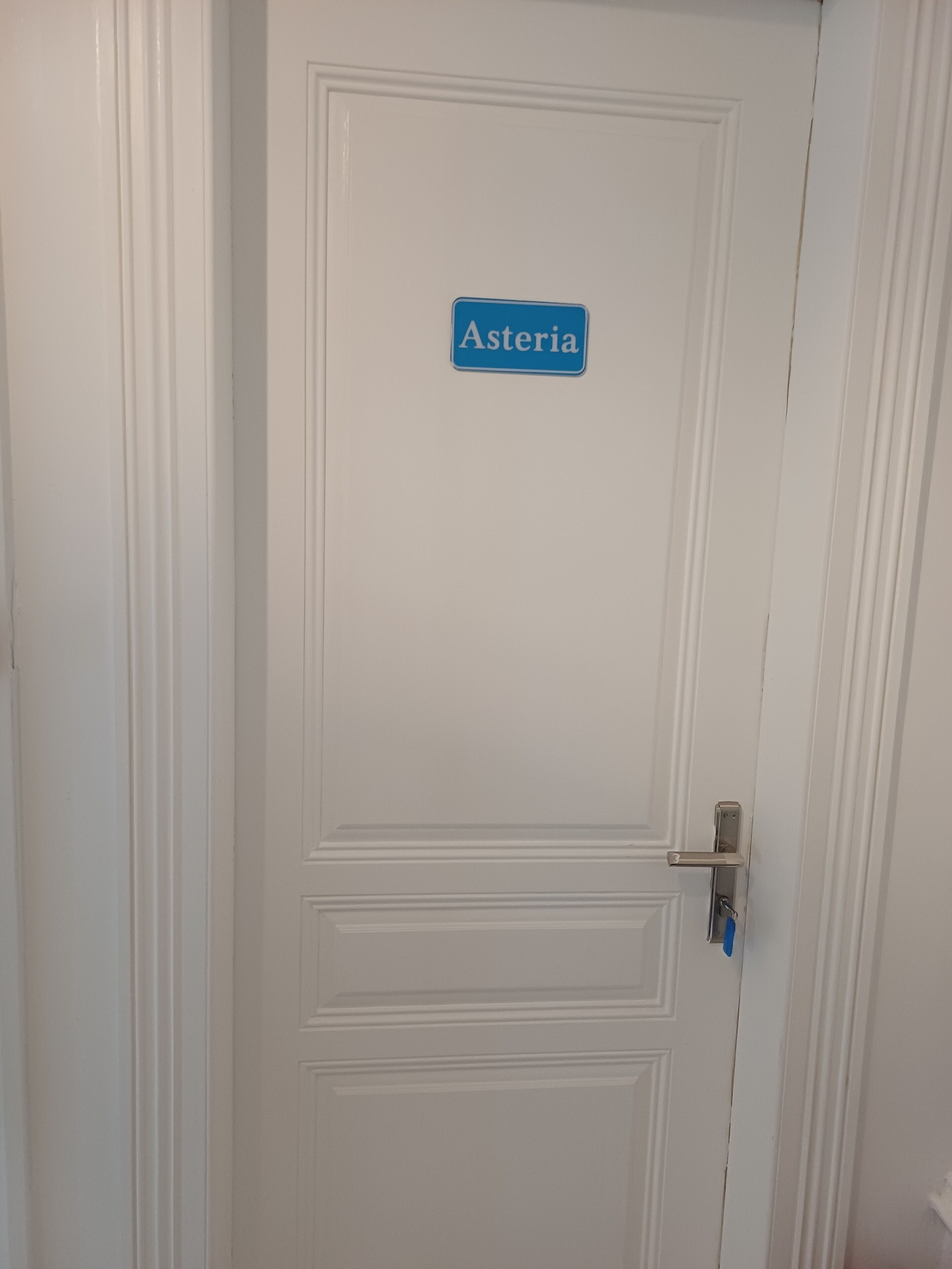 Une porte blanche avec une enseigne bleue portant l’inscription « Asteria » et une serrure à clé.