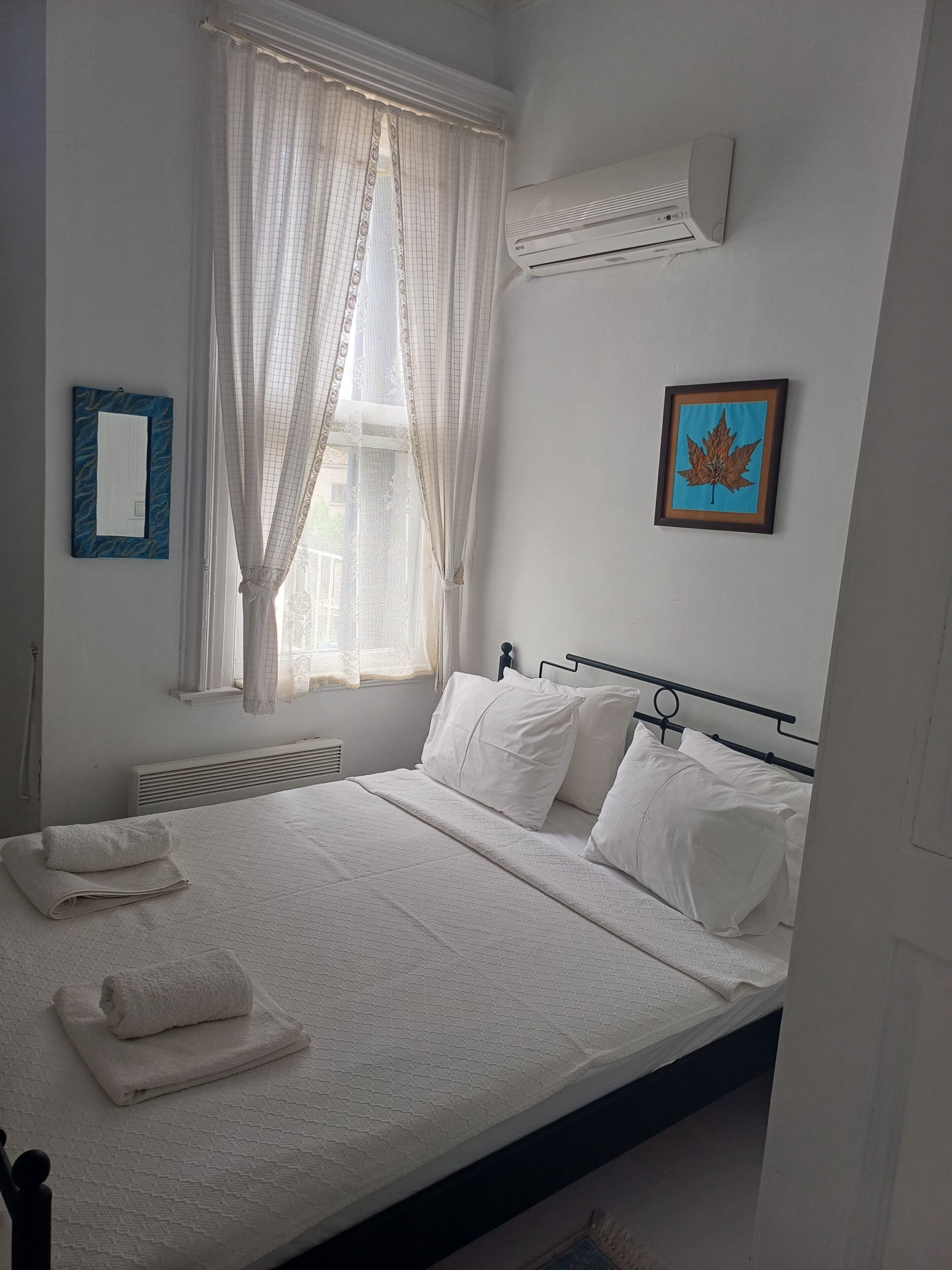 Een netjes opgemaakt bed met witte lakens, een raam met dunne gordijnen, een wandairconditioner, een gelamineerd kunstwerk van een blad en een spiegel met blauwe lijst.