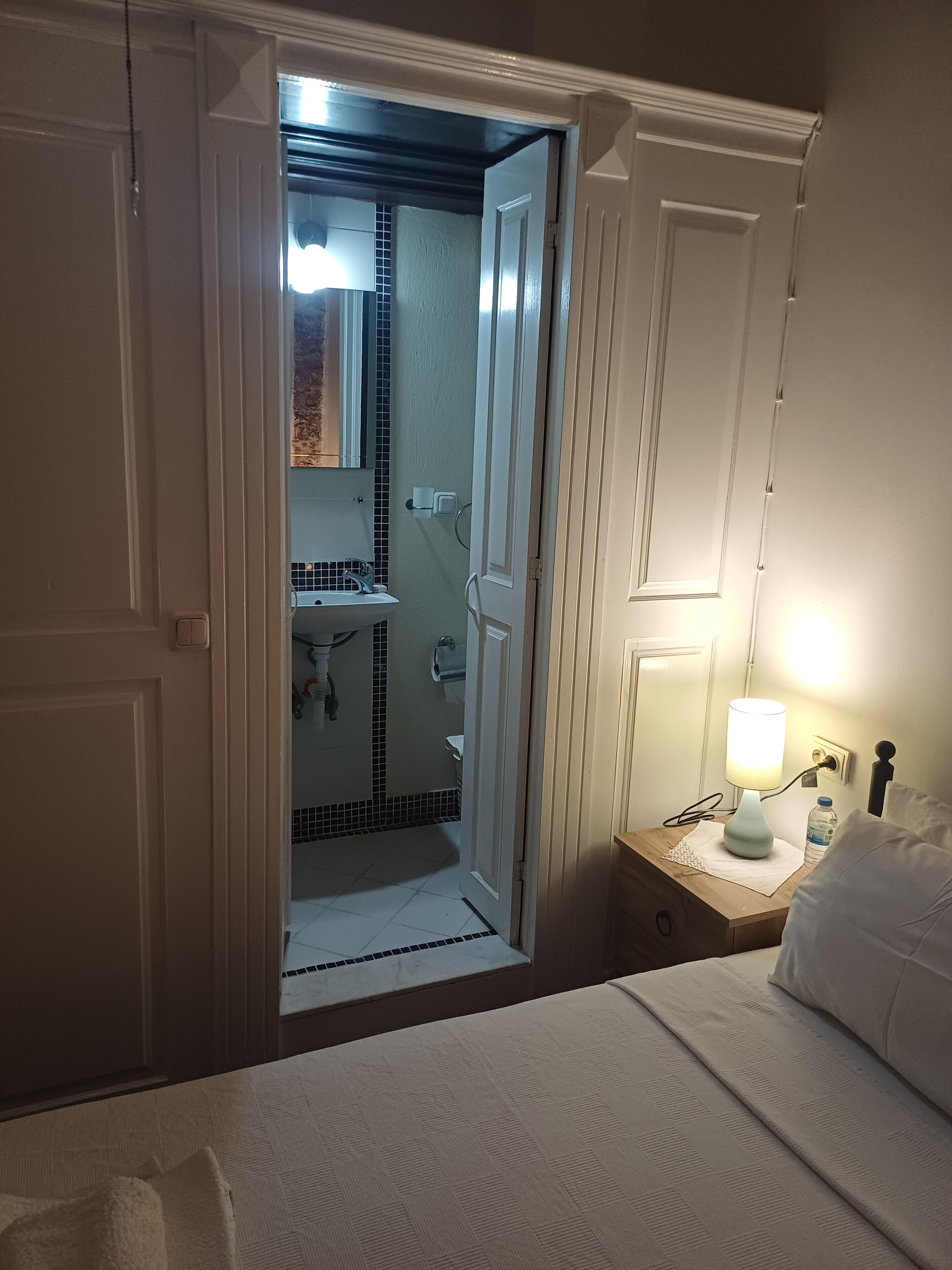 A bedroom with a bed, a bedside table with a lamp, and an open door leading to a bathroom with a sink and mirror.