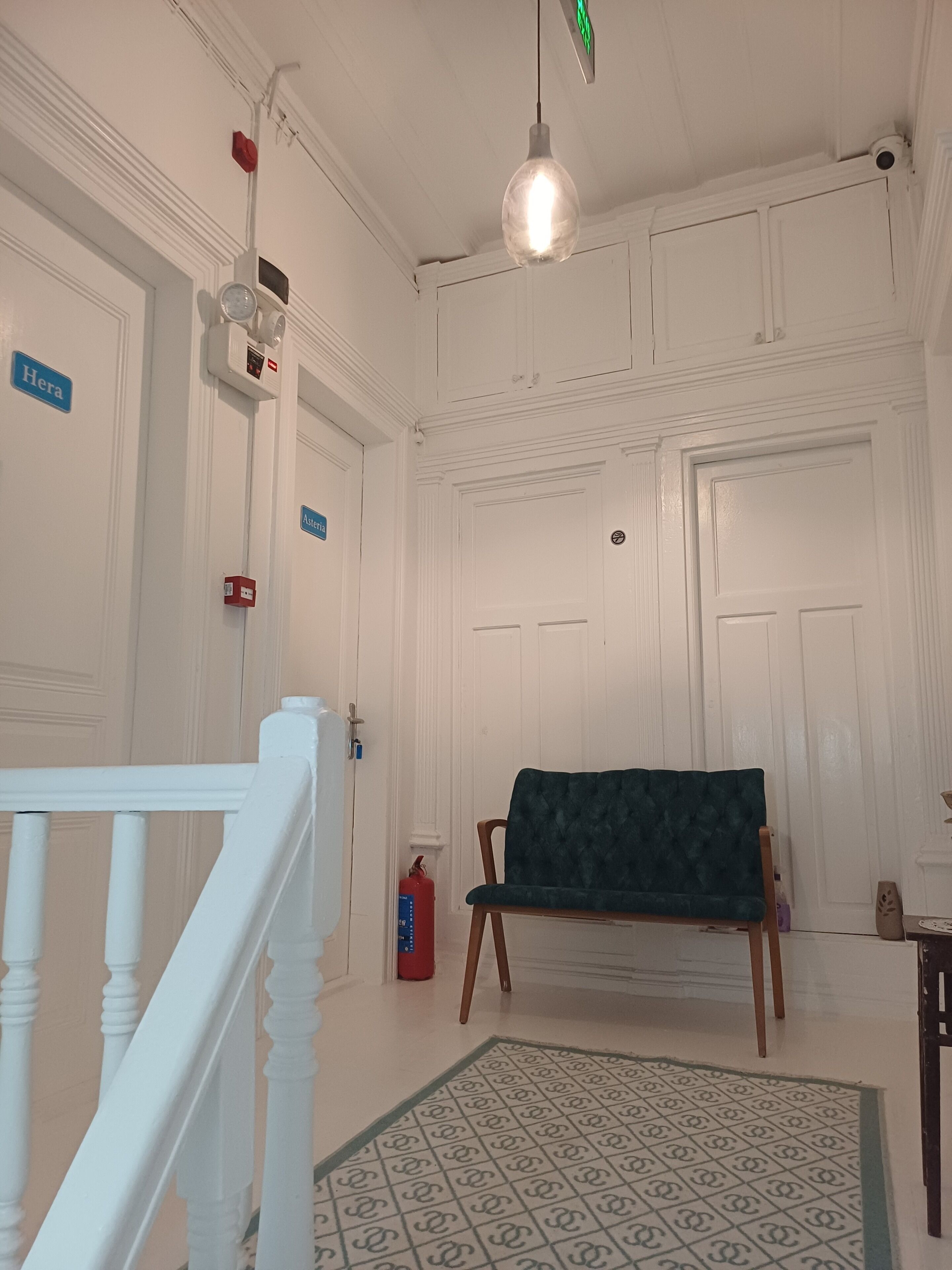 Une pièce aux murs blancs, avec un banc recouvert d’un tissu vert, un tapis à motifs et un escalier blanc.