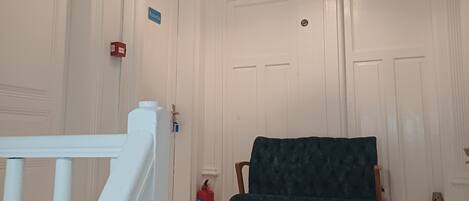 Une pièce aux murs blancs, avec un banc recouvert d’un tissu vert, un tapis à motifs et un escalier blanc.