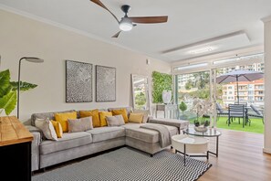 Ein modernes Wohnzimmer mit einem grauen Ecksofa, einem Deckenventilator und Blick auf einen Balkon mit Gartenmöbeln.