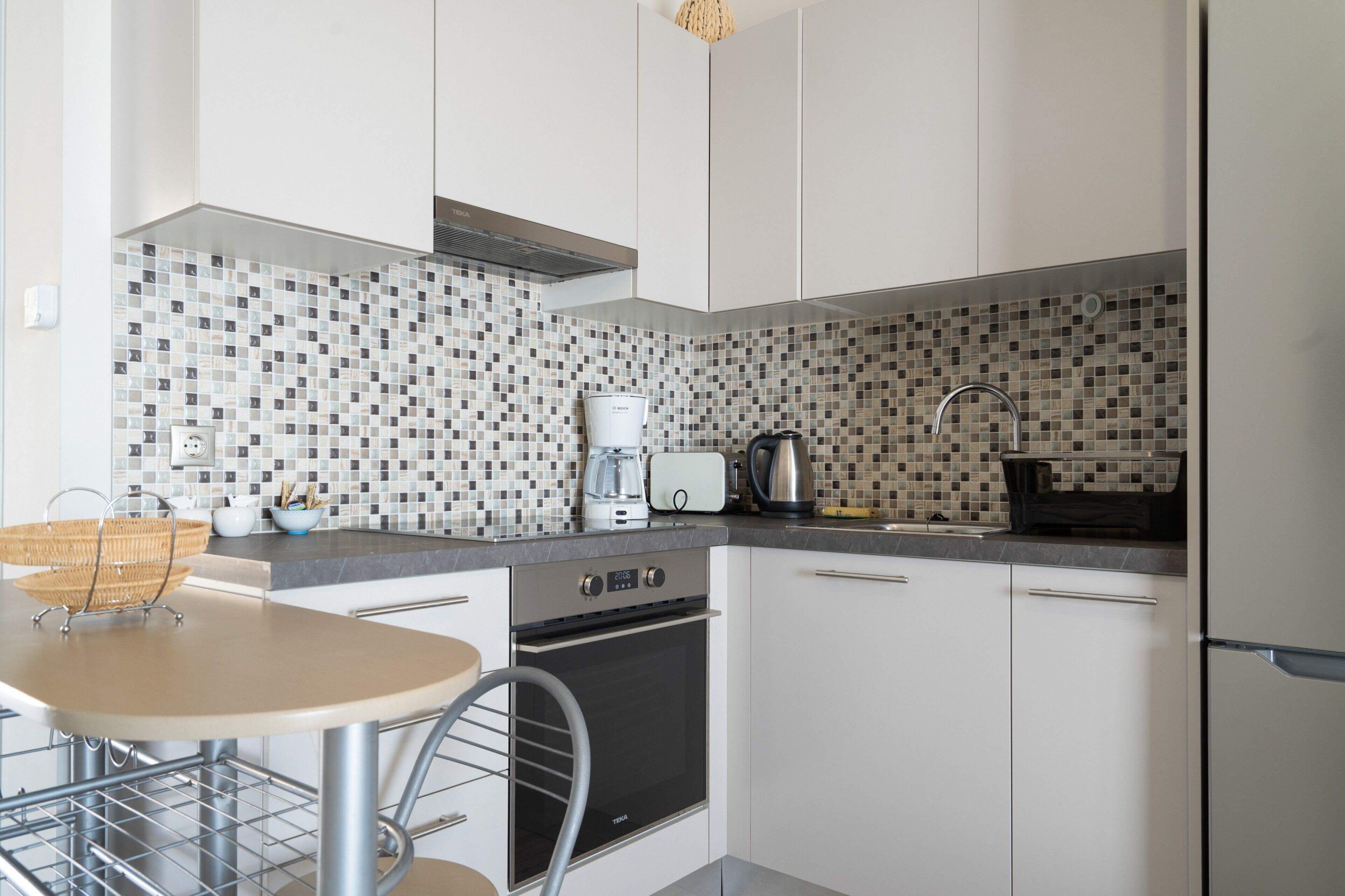 Een moderne keuken met witte kasten, een betegelde spatwand, een roestvrijstalen afzuigkap, een koffiezetapparaat en een broodrooster.