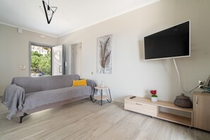 A modern living room with a sofa, a small side table, a TV mounted on the wall, and a view through an open door. - Seaside Retreat with Stunning Sea Views in Kefalonia (Vlachata)