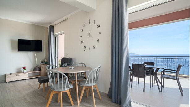 A modern living room with a dining area, a flat-screen TV, a clock, and a balcony with a view.