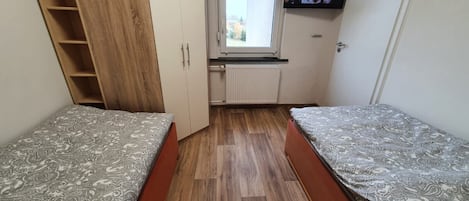 A small room with two beds, a wooden wardrobe, a radiator, a window, and a TV on the wall.
