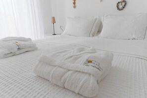 A neatly made bed with white bedding, a folded towel with a logo, and a bedside lamp. - On The Rocks - The Retreat - Serviced Studio (Ios)