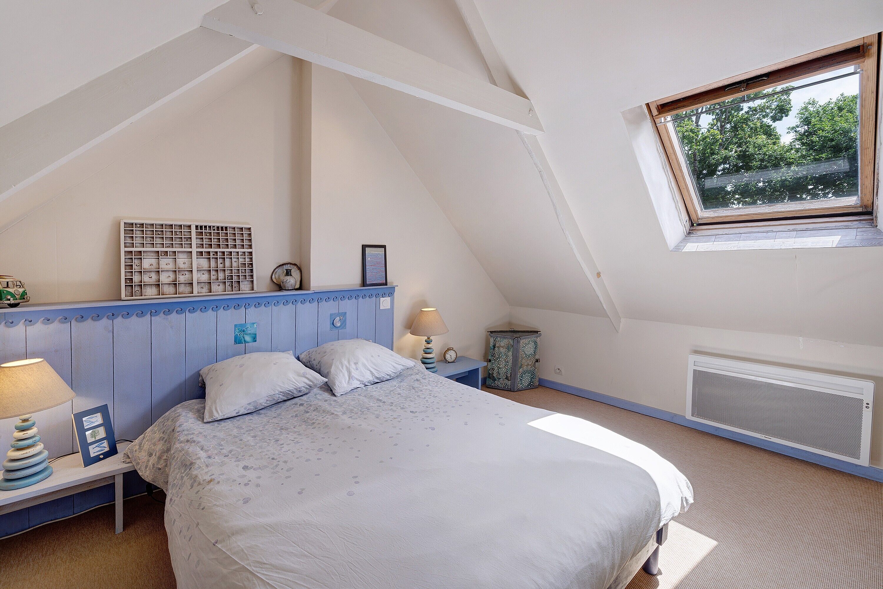 Une chambre avec un plafond en pente, un lit, une table de chevet, une lampe, un tableau encadré, une petite table et une fenêtre donnant sur des arbres.