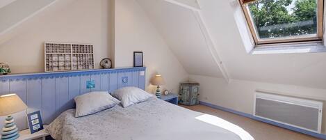 Une chambre avec un plafond en pente, un lit, une table de chevet, une lampe, un tableau encadré, une petite table et une fenêtre donnant sur des arbres.