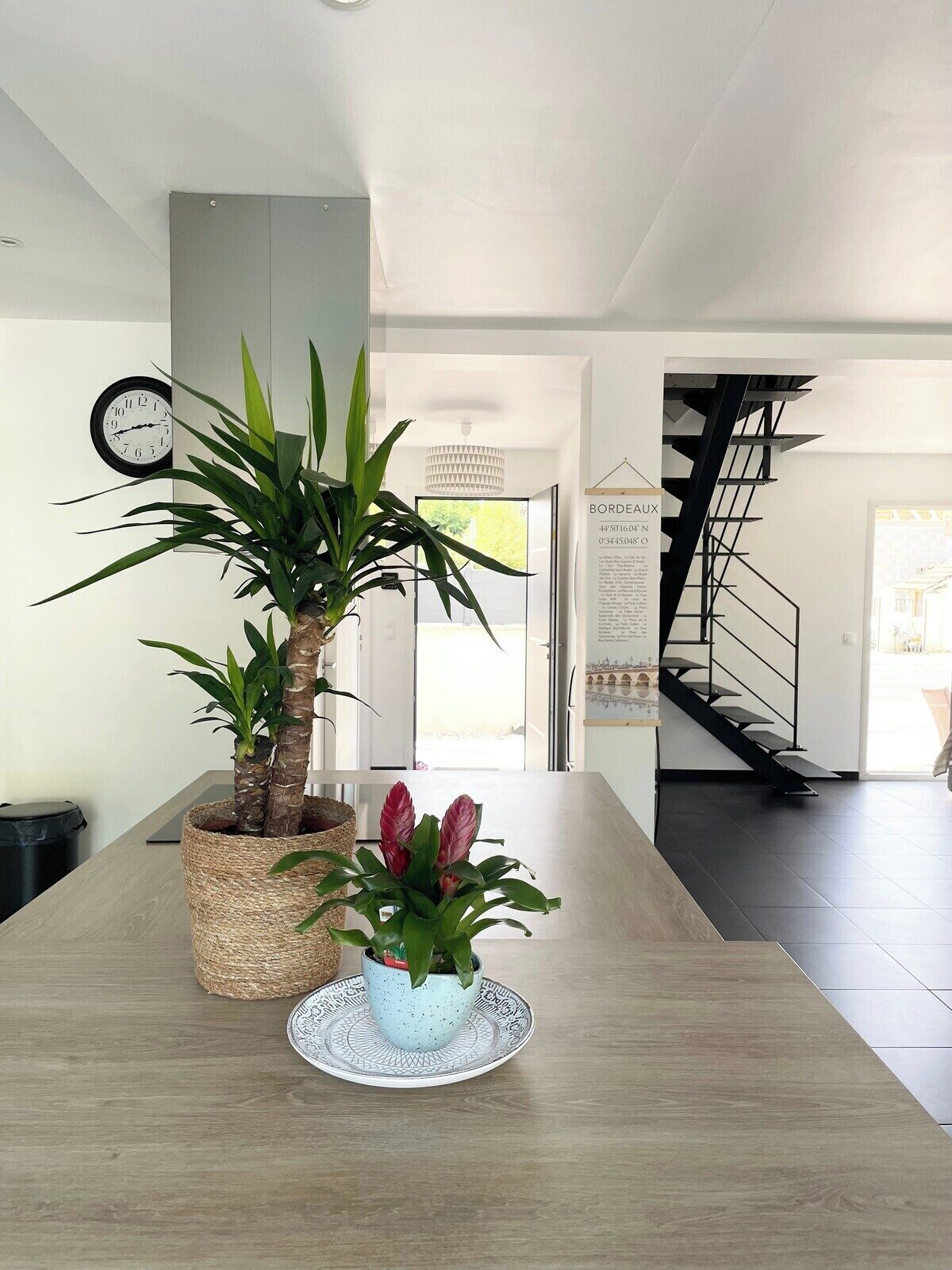 Een modern interieur met een houten tafel, twee planten in potten, een wandklok en een trap.