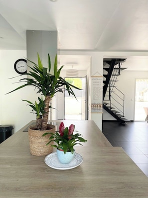 A modern interior with a wooden table, two potted plants, a wall clock, and a staircase.