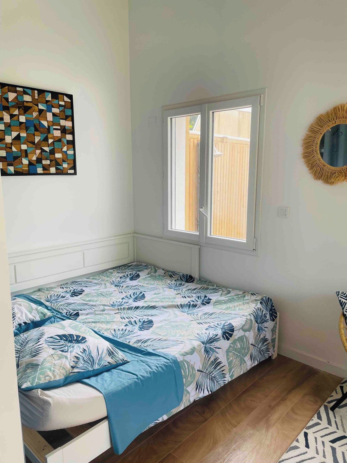 Ein kleines Zimmer mit einem Einzelbett, einem Fenster, einem Wandspiegel und einem geometrischen Kunstwerk.