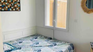 Ein kleines Zimmer mit einem Einzelbett, einem Fenster, einem Wandspiegel und einem geometrischen Kunstwerk.
