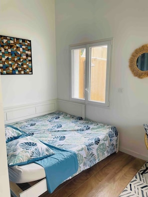 Ein kleines Zimmer mit einem Einzelbett, einem Fenster, einem Wandspiegel und einem geometrischen Kunstwerk.