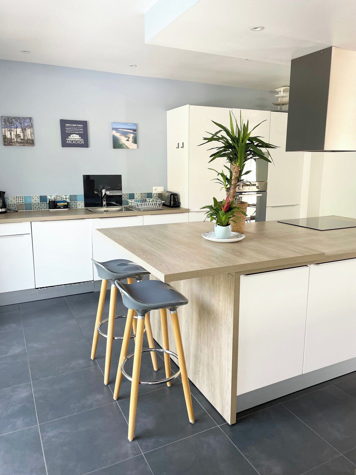 A modern kitchen with a central island, bar stools, a potted plant, and a dining area.