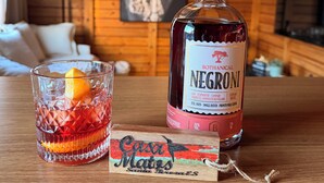 Una botella de ginebra Negroni, un vaso con una rodaja de naranja y un posavasos con el texto 'Ciao Matos' sobre una mesa de madera en una habitación tipo cabaña.