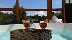 Un jacuzzi con una cesta de mimbre que contiene dos botellas y dos vasos de una bebida roja, rodeado por una terraza de madera y una vista despejada del cielo y los árboles.