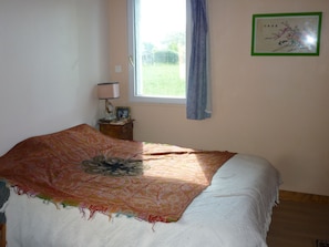 Ein Schlafzimmer mit Bett, Fenster mit Vorhang, Nachttisch mit Lampe und einem gerahmten Bild an der Wand.