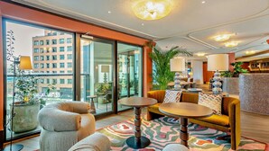 En moderne hotellobby med en stor glasdør, der fører til en balkon, et siddeområde med sofaer og borde, samt en dekorativ lampe.