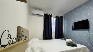 Een slaapkamer met een bed, een nachtkastje, een wandtelevisie en een raam met gordijnen.
