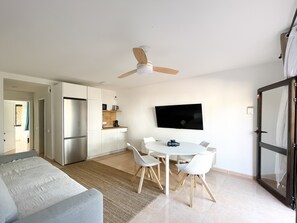 Un salon moderne comprenant un canapé, une table à manger, des chaises, un ventilateur de plafond, un téléviseur et un coin cuisine équipé d’appareils électroménagers.