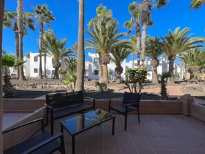 A patio with outdoor seating, palm trees, and white buildings in the background. - Oasis Duna Sand & Soul – Stylish 5-Person Apartment with Terrace and Pool (Corralejo)