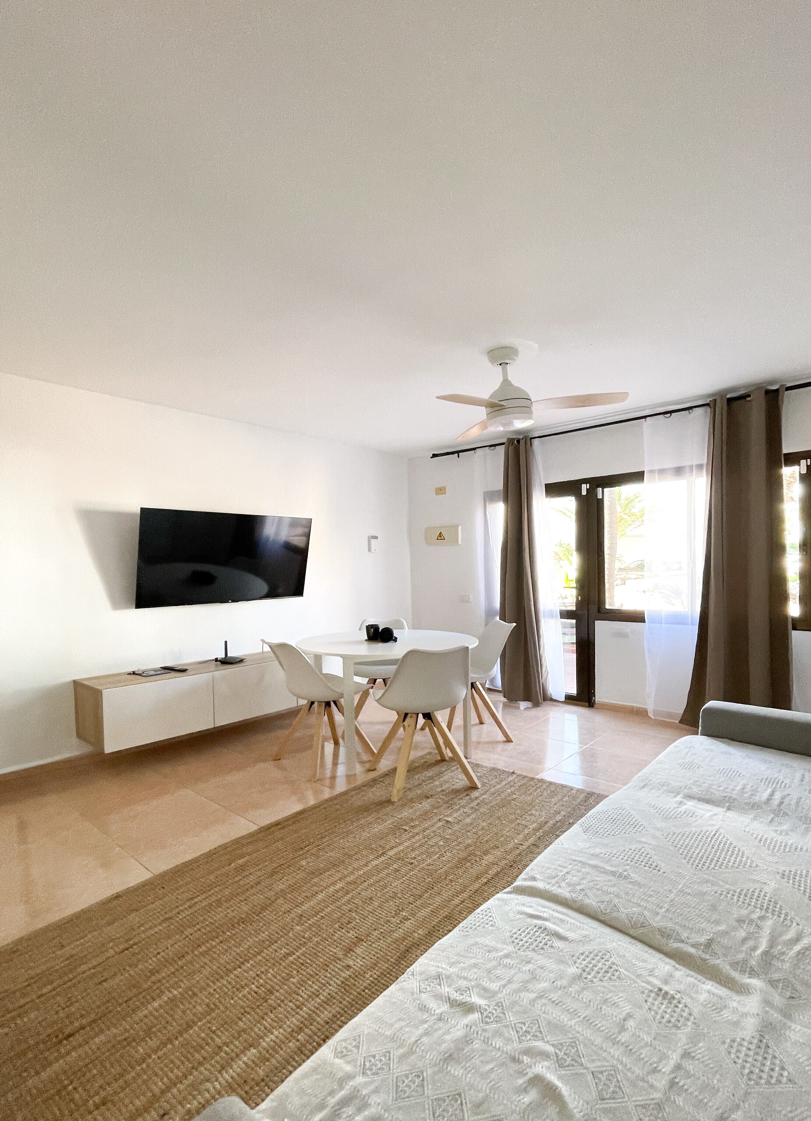 A modern hotel room with a flat-screen TV, a dining table with chairs, a ceiling fan, and a large bed.