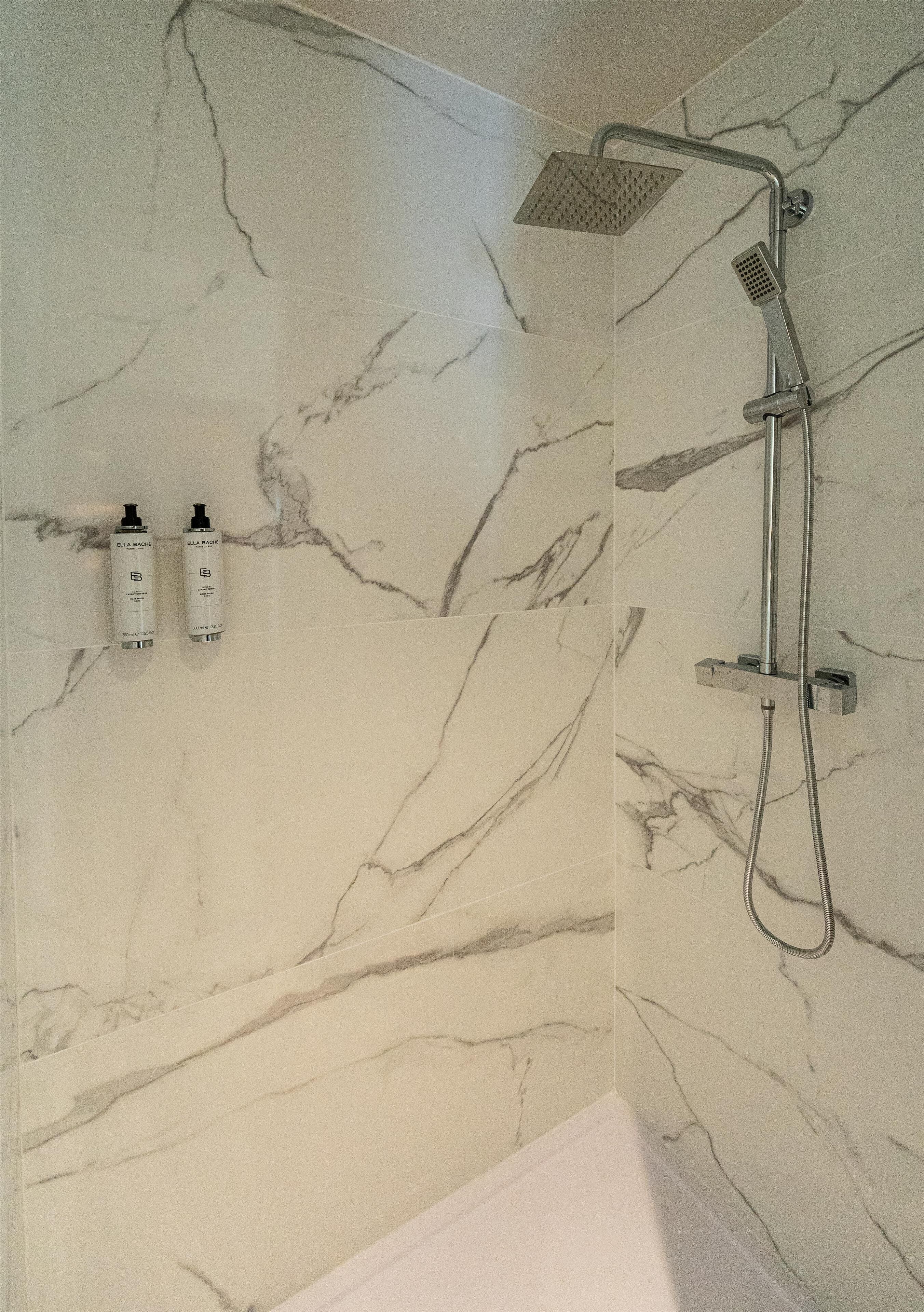 Un baño con pared de mármol, ducha con cabezal de ducha manual y dos botellas de champú.