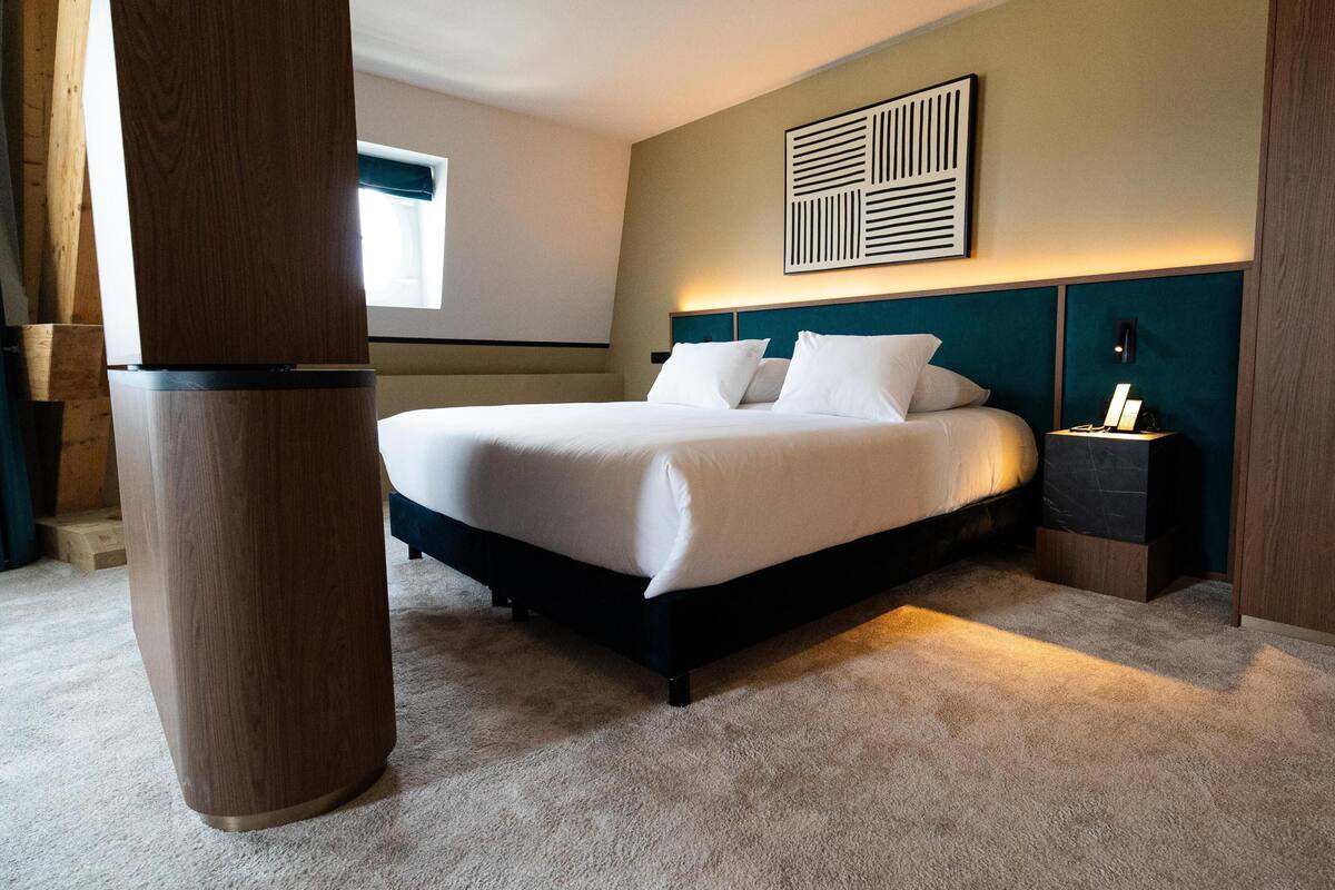 Una habitación de hotel con cama, mesita de noche, una obra de arte en la pared y una ventana.