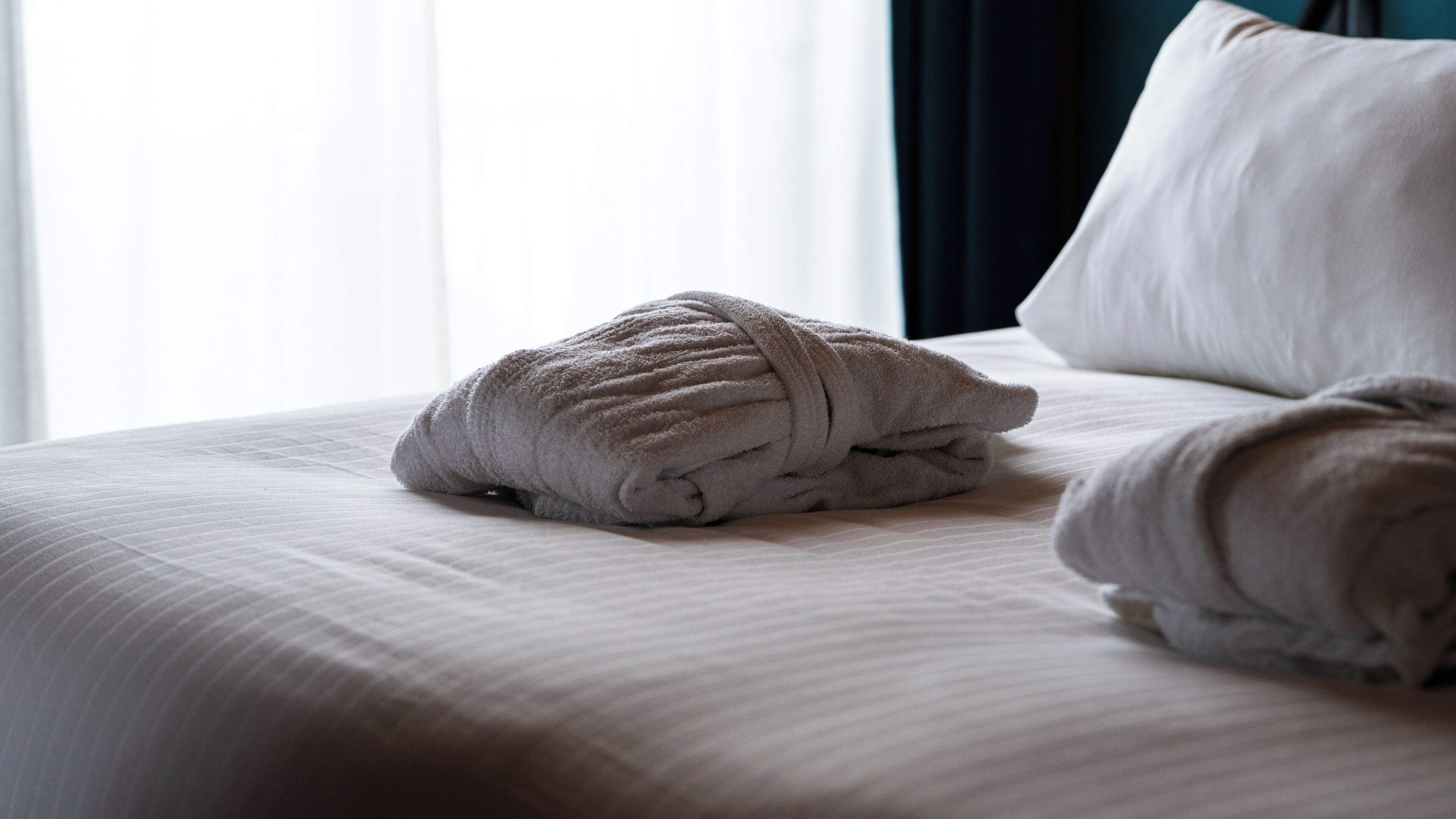 Una cama bien hecha con una toalla enrollada encima, una almohada y una ventana con cortinas.