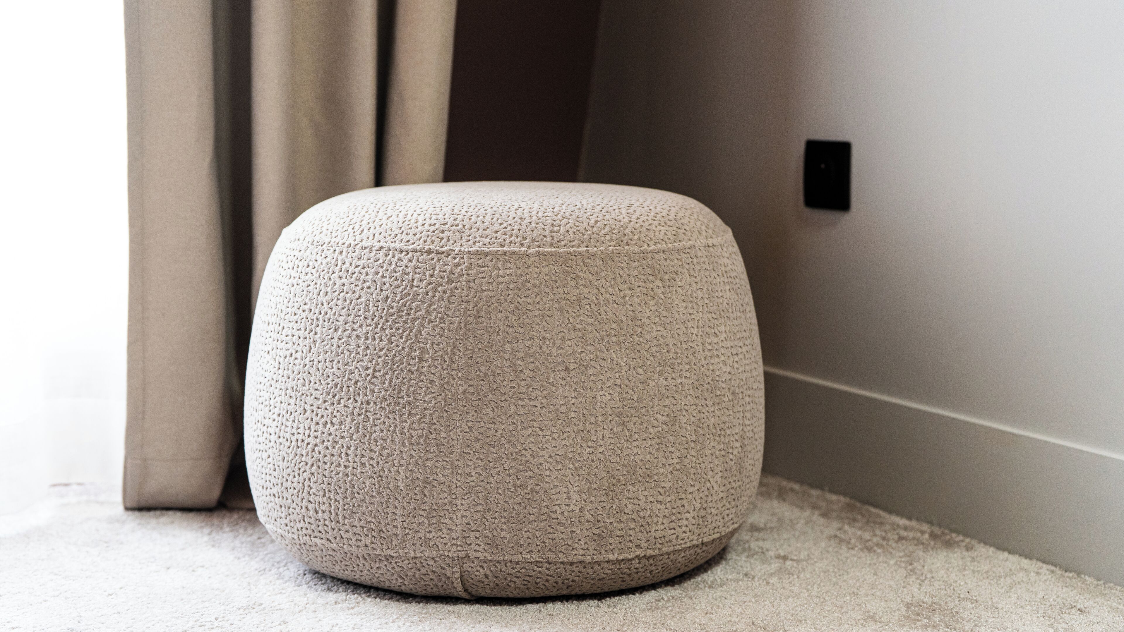 A round, textured ottoman in a neutral color, placed on a carpeted floor against a wall with a switch.